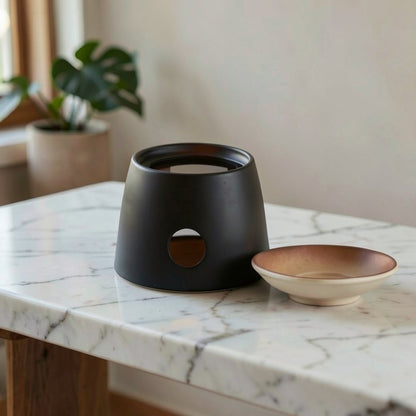 A matte black ceramic candle holder with a circular cutout, displayed on a white marble table next to a small beige and brown ceramic dish in a softly lit indoor setting.
